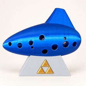 Peut inclure: Un ocarina bleu vif avec plusieurs trous pour les doigts, reposant sur un support trapézoïdal gris clair. Le support présente un emblème triangulaire doré composé de trois petits triangles disposés en forme de triangle.