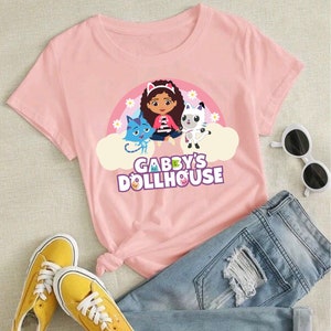May include: Pink t-shirt featuring a graphic of Gabby from Gabby's Dollhouse, a cartoon character with brown hair and a pink headband, sitting on a cloud with two cats. The text "Gabby's Dollhouse" is printed below the graphic.