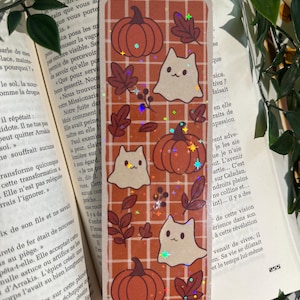 Peut inclure: Marque-page rectangulaire sur le thème de l'automne. Il présente des citrouilles oranges, des feuilles marron et des chats fantômes blancs sur un fond à carreaux. Le marque-page a un effet de paillettes holographiques.