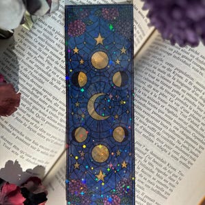 Peut inclure: Marque-page rectangulaire avec un motif céleste. Il présente un fond bleu foncé avec des étoiles dorées, des croissants de lune et des phases de la lune. Des accents floraux violets et du texte sont également visibles.