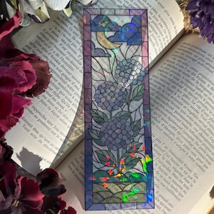 Peut inclure: Un signet rectangulaire avec un motif de vitrail représentant des hortensias bleus, un croissant de lune et des nuages. Le signet a une finition holographique irisée et est posé sur un livre ouvert.