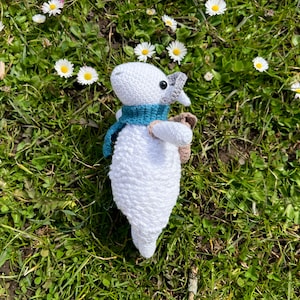 May include: White crocheted lamb toy with a blue scarf and a brown backpack, sitting in a field of green grass and white daisies.