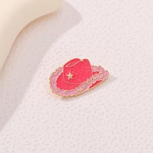 May include: A pink cowboy hat enamel pin with a gold outline and a star detail. The hat has a fluffy, light pink trim. This accessory is perfect for adding a touch of Western flair to any outfit.