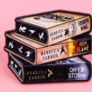 May include: A stack of book-shaped pins, each displaying a different title: "Fourth Wing", "Iron Flame", and "Onyx Storm" by Rebecca Yarros. The pins have black spines and tan covers with text and illustrations. Set against a pink backdrop.