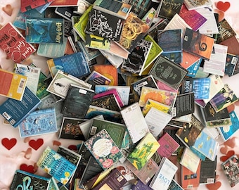 May include: A large pile of paperback books with various titles and authors. The books are arranged in a random order and some are stacked on top of each other. The books are all different colors and sizes. The books are on a pink surface with red hearts scattered around them.
