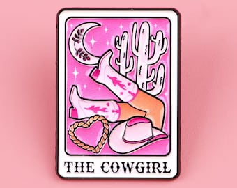 May include: A pink and white graphic featuring a cowgirl theme. The design includes a crescent moon, cactus, cowboy boots, a heart-shaped rope, and a cowboy hat. The text "THE COWGIRL" is at the bottom of the design.