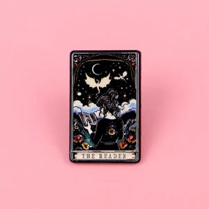 May include: A rectangular pin badge with a tarot card design. It depicts a woman reading cards beneath a night sky with a crescent moon and stars. The pin has a black border and reads "THE READER".