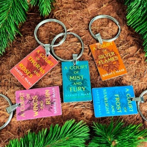 May include: A collection of keyrings with rectangular tags displaying book covers from Sarah J. Maas's "A Court of Thorns and Roses" series. Each tag features a book title and author's name, attached to a silver keyring. The keyrings are in various colors.