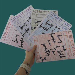 May include: A hand displays a collection of library cards in pastel colours. Each card features the words "LIBRARY CARD" and handwritten phrases such as "buy me books" and "buy me & tell I'm pretty".
