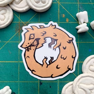 May include: A die-cut sticker featuring a tan, cartoon-style creature curled into a circular shape, biting its own tail. The creature has dark brown outlines, sharp teeth, and small limbs. The sticker is surrounded by several white, circular clay tokens.