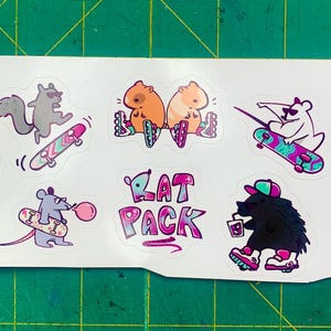 Puede incluir: Una hoja de pegatinas con varios animales haciendo skate y sosteniendo bebidas. Las pegatinas son coloridas y presentan un degradado rosa y azul con el texto "Rat Pack" en un degradado similar.