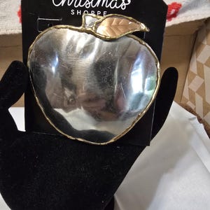 May include: A silver-colored apple-shaped ornament with a gold-colored rim and a copper-colored leaf. The ornament is on a black tag that reads "The Christmas Shoppe" in white script.