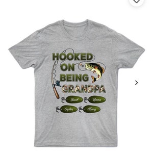 May include: Gray t-shirt with a green camouflage design and the text "Hooked on being Grandpa" with a fishing rod and fish graphic. The names Jacob, Emma, Sophia, and Henry are on fishing lure tags.