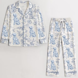 May include: A matching pajama set featuring a button-down shirt and pants. The set is cream-colored with a blue floral and dog pattern. The shirt has a notched collar and blue trim. The pants have a drawstring waist and blue trim.