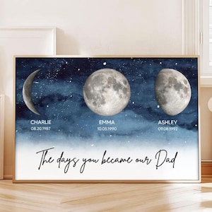 Op de afbeelding: Een ingelijste print met drie maanfasen tegen een donkerblauwe aquarelachtergrond met sterren. Namen en data staan onder elke maanfase, met de tekst "The days you became our Dad" onderaan.