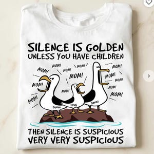 Può includere: T-shirt bianco con una grafica umoristica. Gabbiani dei cartoni animati con fumetti che dicono "MOM!" e nomi. Il testo dice "SILENCE IS GOLDEN UNLESS YOU HAVE CHILDREN" e "THEN SILENCE IS SUSPICIOUS VERY VERY SUSPICIOUS."