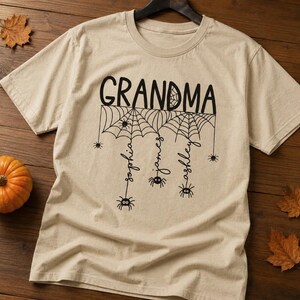 May include: A beige short-sleeve t-shirt featuring a black graphic of spider webs and spiders. The text "GRANDMA" is printed at the top, with the names "sophia", "james", and "ashley" hanging from the webs, each ending in a small spider.