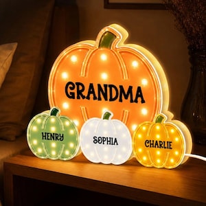 May include: A large orange pumpkin-shaped light with the word GRANDMA in black text, accompanied by three smaller pumpkin-shaped lights in green, white, and yellow, featuring the names HENRY, SOPHIA, and CHARLIE in black text respectively.