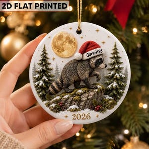 May include: A round white ceramic Christmas ornament featuring a raccoon wearing a red Santa hat with the name Jimothy printed on it. The design includes a full moon, evergreen trees, snow, and the year 2026. It hangs from a gold string.