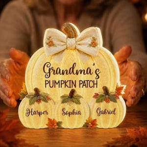 May include: A light-colored pumpkin-shaped decorative sign featuring a large bow at the top and the text Grandma's PUMPKIN PATCH. Three smaller pumpkins are attached at the base, each inscribed with the names Harper, Sophia, and Gabriel.