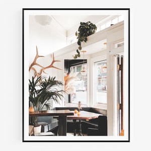 May include: A white-walled cafe interior with a wooden table and chairs. A large potted plant and antlers are on the wall. A window with a view of the street is in the background.