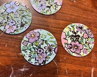 May include: Four round coasters with floral designs. Each coaster features a different arrangement of purple, pink, and white flowers with green leaves on a light blue background. Gold accents are scattered throughout the floral patterns.