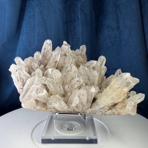 May include: A large cluster of clear, pointed quartz crystals with natural inclusions and varying heights. The mineral specimen is mounted on a square, transparent acrylic stand with a silver-toned metal support frame for display purposes.