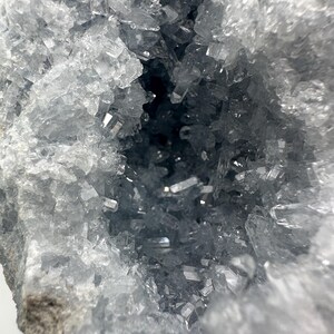 May include: A close-up of a geode featuring a cluster of light blue celestite crystals. The translucent crystals have a sharp, geometric shape. The geode's interior is a deep, dark blue.