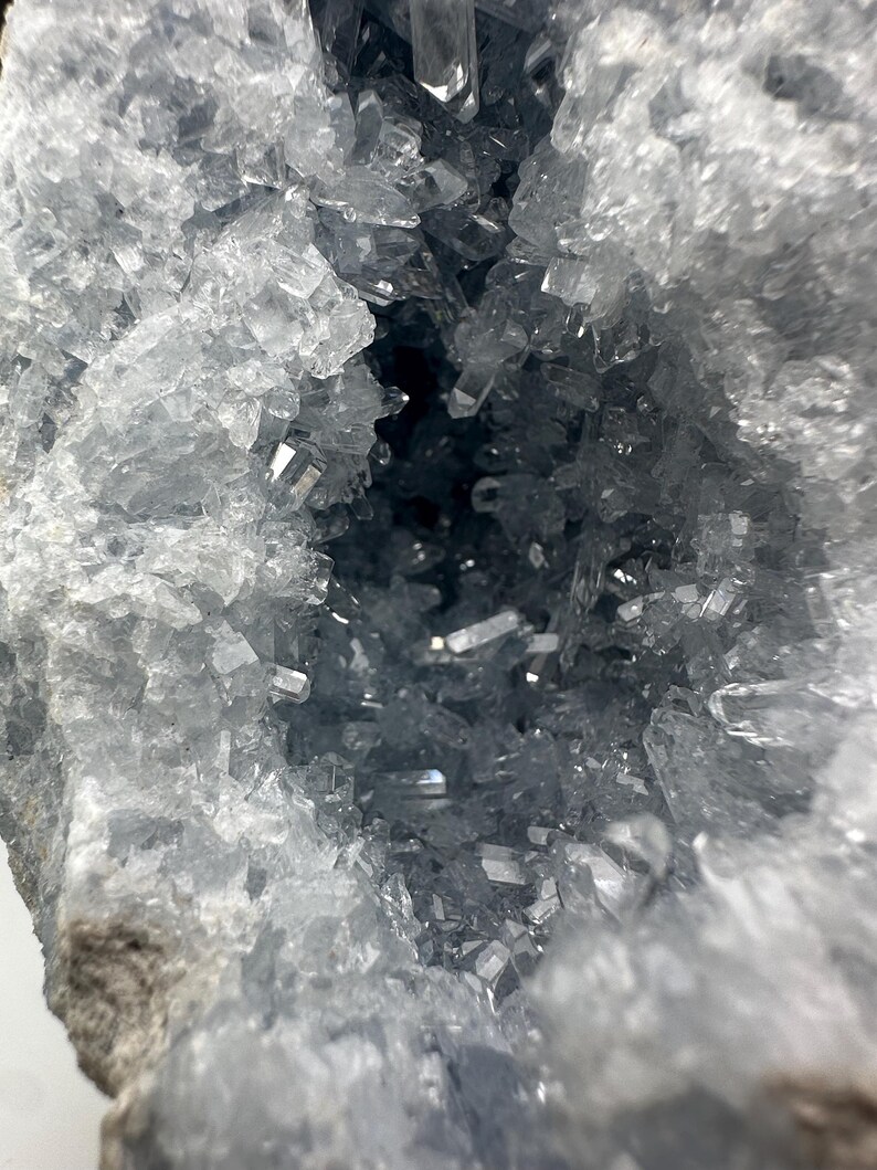 May include: A close-up of a geode featuring a cluster of light blue celestite crystals. The translucent crystals have a sharp, geometric shape. The geode's interior is a deep, dark blue.