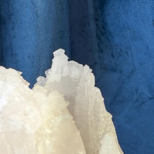 May include: A translucent white mineral specimen with jagged, crystalline edges and a rough, textured surface. The structure features sharp, pointed peaks and layered formations that catch the light, creating a stark contrast against the deep blue backdrop.