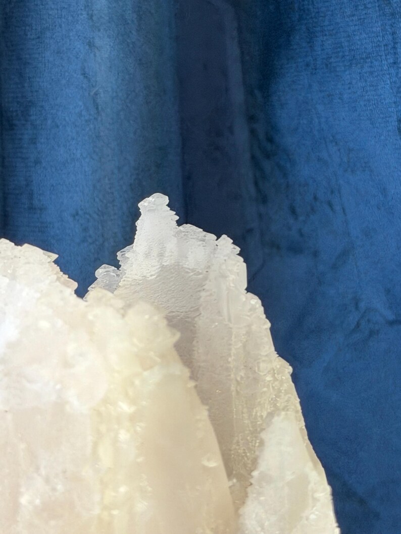 May include: A translucent white mineral specimen with jagged, crystalline edges and a rough, textured surface. The structure features sharp, pointed peaks and layered formations that catch the light, creating a stark contrast against the deep blue backdrop.
