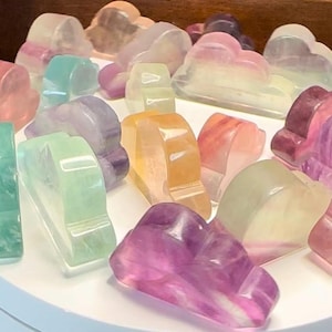 May include: A collection of colorful, translucent cloud-shaped decorative objects. The objects are in shades of pink, purple, green, yellow, and orange. They are arranged on a white surface, creating a visually appealing display.