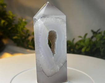 May include: A polished, rectangular crystal with a pointed top. The crystal is primarily gray with a white and light gray interior, featuring a central cavity. The crystal is displayed on a white surface, with greenery in the background.