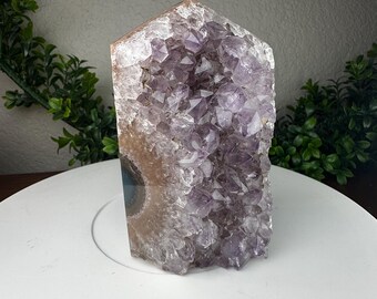 May include: A large amethyst crystal geode with a polished edge. The crystal features a deep purple color with a crystalline structure. The geode is set on a white surface, with green foliage in the background. The geode is a natural home decor item.