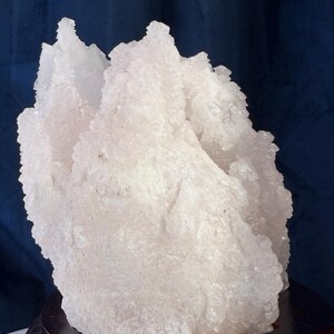 May include: A translucent, pale pink crystalline mineral specimen with a jagged, multi-pointed surface texture. The cluster is mounted on a circular, dark brown polished wooden base, showcasing natural mineral formations and irregular edges.
