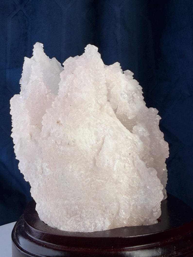 May include: A translucent, pale pink crystalline mineral specimen with a jagged, multi-pointed surface texture. The cluster is mounted on a circular, dark brown polished wooden base, showcasing natural mineral formations and irregular edges.