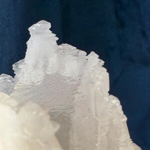 May include: A translucent white mineral specimen with jagged, crystalline formations. The structure features sharp, angular edges and a rough, layered texture that catches the light, creating a stark contrast against the dark blue background.