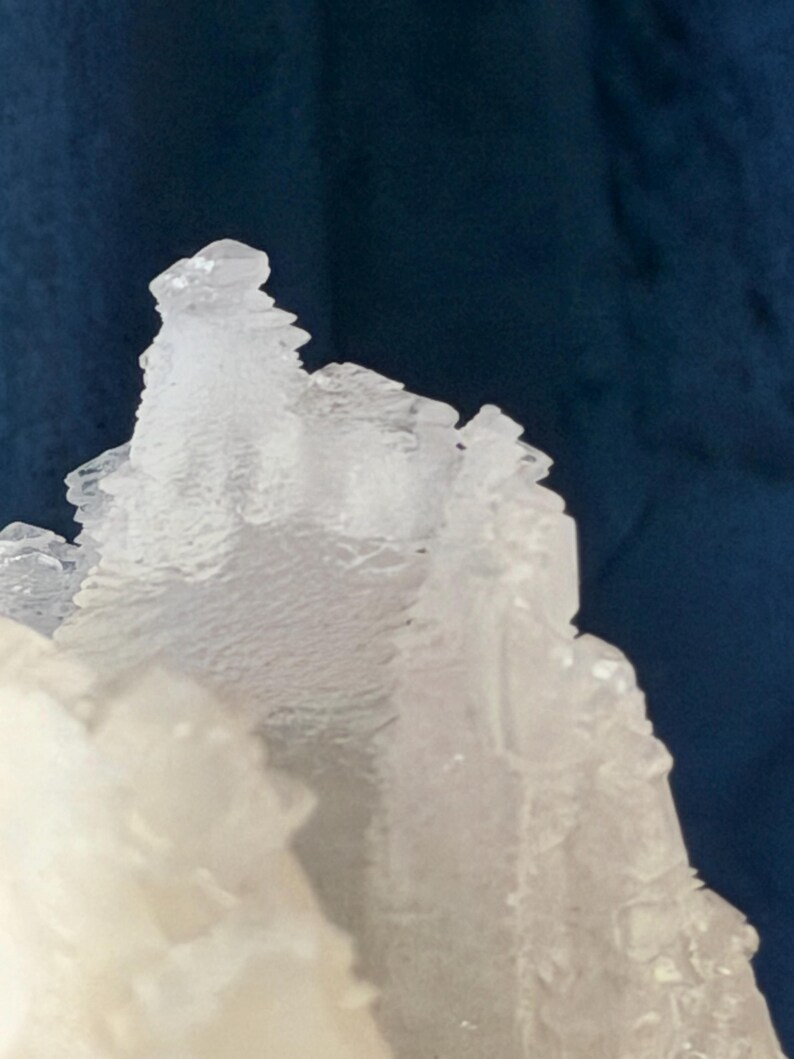 May include: A translucent white mineral specimen with jagged, crystalline formations. The structure features sharp, angular edges and a rough, layered texture that catches the light, creating a stark contrast against the dark blue background.