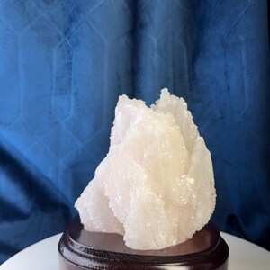 May include: A pale pink, translucent mineral specimen with a jagged, crystalline texture. The cluster features multiple sharp, pointed peaks and irregular facets. It is securely mounted on a smooth, circular, dark brown wooden display base.