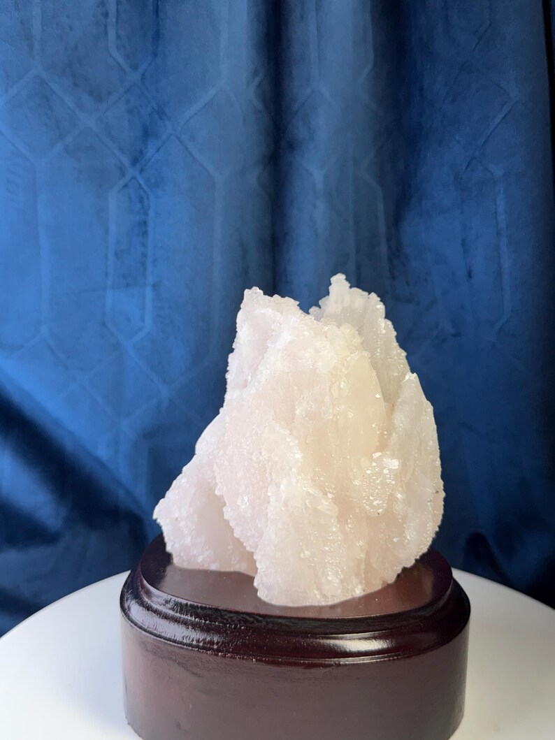 May include: A pale pink, translucent mineral specimen with a jagged, crystalline texture. The cluster features multiple sharp, pointed peaks and irregular facets. It is securely mounted on a smooth, circular, dark brown wooden display base.