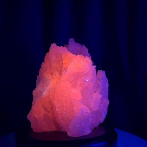 May include: A crystalline mineral specimen exhibiting a vibrant, glowing neon pink fluorescence under ultraviolet light. The cluster features a jagged, irregular surface with numerous small, translucent facets that catch the light, set atop a circular base.