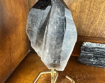 May include: A large, dark grey, double-terminated crystal point on a gold metal hexagonal stand. The crystal has a smoky, translucent appearance.