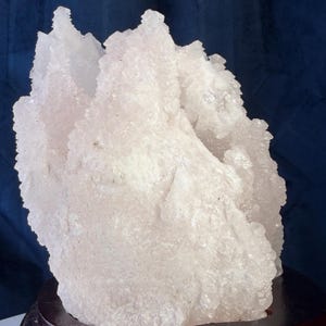 May include: A pale pink, translucent mineral cluster with a jagged, crystalline surface texture. The specimen features multiple pointed peaks and irregular formations, mounted securely on a smooth, circular, dark brown wooden display base.