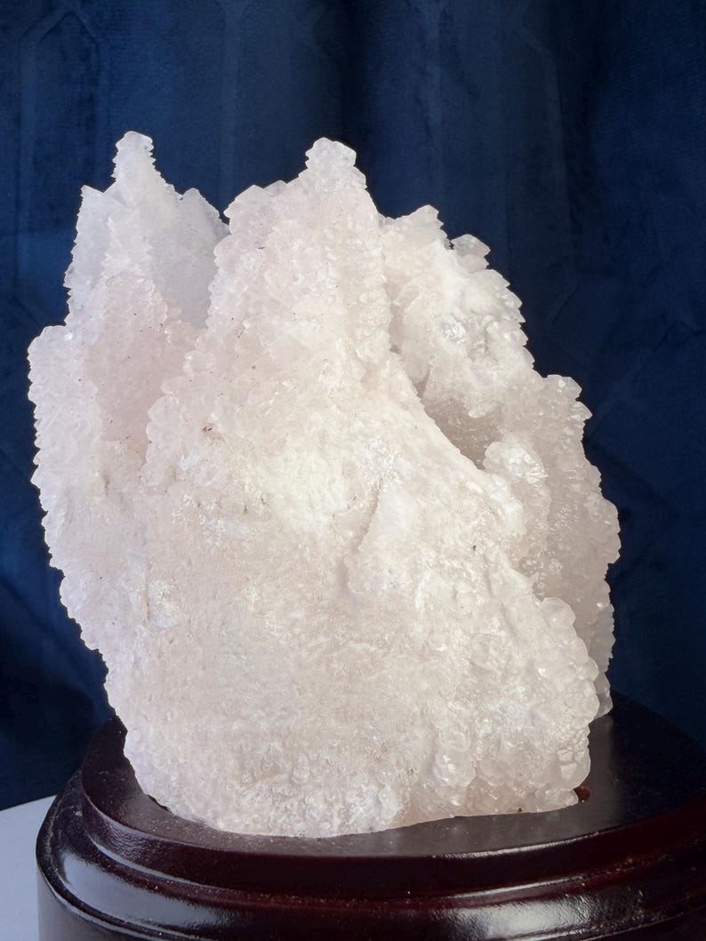 May include: A pale pink, translucent mineral cluster with a jagged, crystalline surface texture. The specimen features multiple pointed peaks and irregular formations, mounted securely on a smooth, circular, dark brown wooden display base.