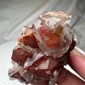 May include: A cluster of translucent, reddish-brown quartz crystals. The crystals have a faceted, geometric structure, with varying shades of red and brown. The overall shape is irregular and natural, with a rough texture and a mix of clear and opaque areas.