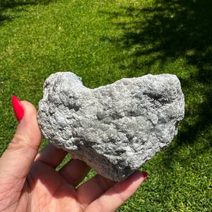 May include: A grey, heart-shaped rock with a rough, textured surface. The rock is held in a hand with red nail polish, set against a backdrop of green grass and sunlight.