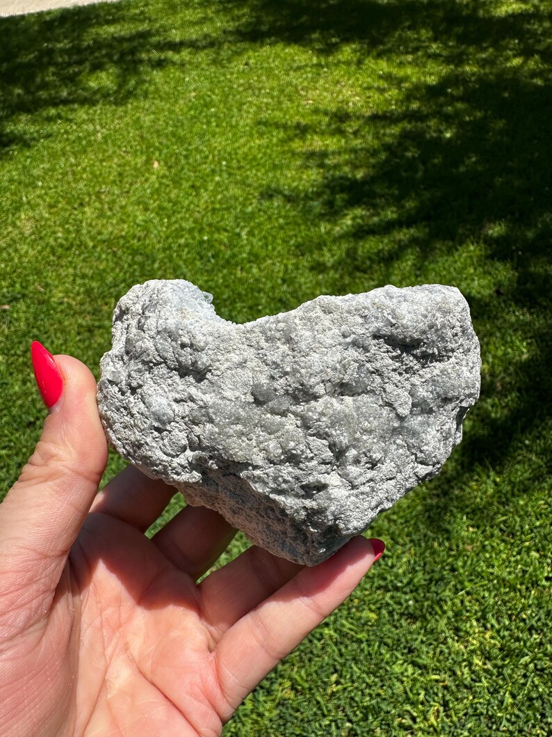 May include: A grey, heart-shaped rock with a rough, textured surface. The rock is held in a hand with red nail polish, set against a backdrop of green grass and sunlight.
