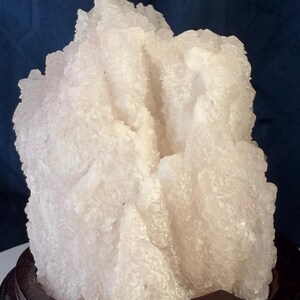 May include: A large, translucent white crystal cluster with jagged, multifaceted surfaces and a rough, uneven texture. The mineral specimen is mounted on a smooth, dark brown, circular wooden base, showcasing a natural, irregular formation.