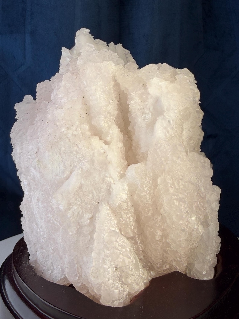May include: A large, translucent white crystal cluster with jagged, multifaceted surfaces and a rough, uneven texture. The mineral specimen is mounted on a smooth, dark brown, circular wooden base, showcasing a natural, irregular formation.