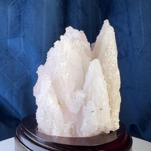 May include: A cluster of translucent white quartz crystals with jagged, pointed edges and a textured, crystalline surface. The mineral formation is securely mounted on a smooth, circular, dark brown wooden base with a polished finish.